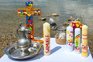 Taufschale, Taufkerzen mit Namen, buntes Kreuz, Wasser eines Sees im Hintergrund